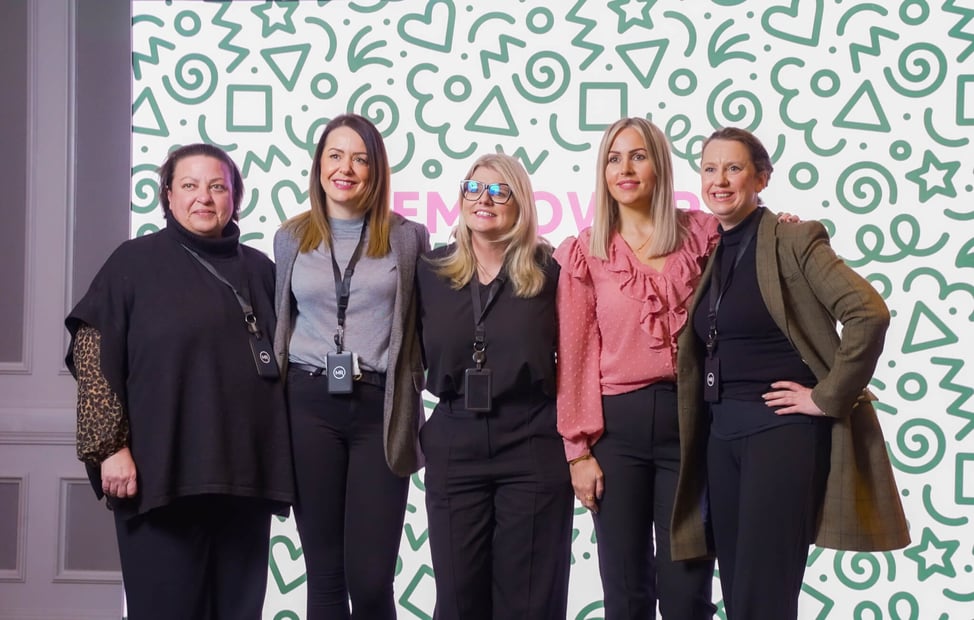 "Five women from the Map Room team standing side by side and smiling in front of a patterned backdrop with green doodles and the partially visible word ‘EMPOWER’."